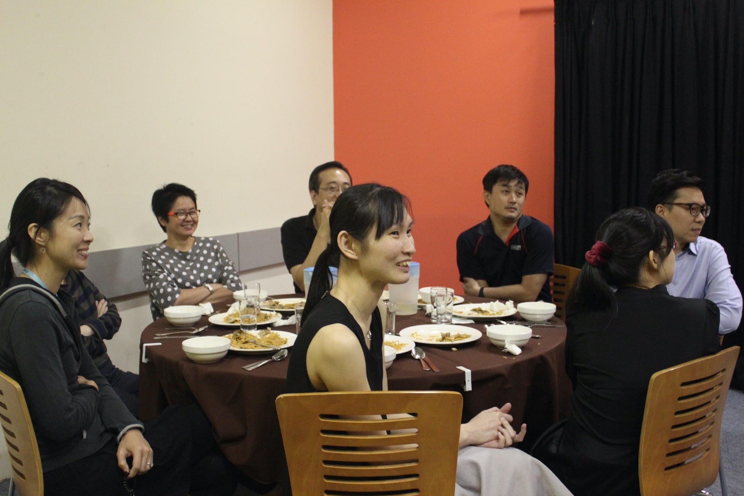 Seven people sit around a round table with food and drinks, smiling and conversing in a room with an orange wall.